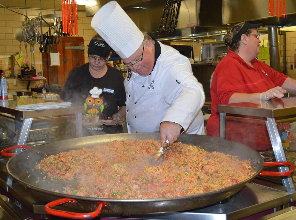 CHEF BRIAN ADDS all batches of the paella into the patella to keep hot until serving. At back left is Johannah Holland of the kitchen staff, and at right, Carol Kipfer, also of the kitchen staff.